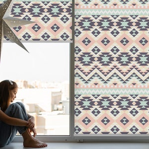 Könnte beinhalten: Ein Fenster mit einem gemusterten Rollo mit einem geometrischen Design in Rosa-, Grün- und Grautönen. Eine junge Person sitzt auf der Fensterbank und schaut aus dem Fenster.