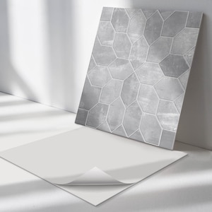May include: A sheet of peel and stick tile backsplash with a geometric pattern of gray hexagonal tiles. The sheet is laying on a white surface with a corner peeled back to show the adhesive backing.