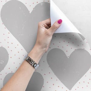 May include: A white background with a repeating pattern of gray hearts and small pink dots. A hand is peeling back a corner of the wallpaper to reveal the pattern.