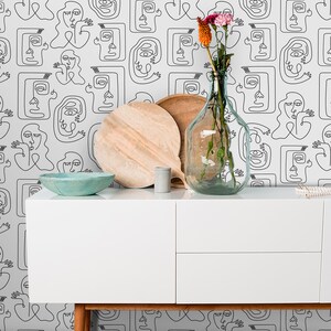 K&ouml;nnte beinhalten: Eine wei&szlig;e Kommode mit zwei Schubladen steht vor einer Wand, die mit einer schwarz-wei&szlig;en Linienkunst-Tapete mit abstrakten Gesichtern bedeckt ist. Auf der Kommode stehen ein Holzbrett, ein kleiner grauer Keramikbeh&auml;lter und eine klare Glasvase mit getrockneten Blumen.