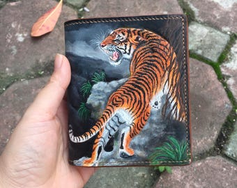 May include: A brown leather wallet with a hand-painted image of a tiger in a jungle setting. The tiger is orange and black with white stripes. The wallet has a dark brown background and green foliage.