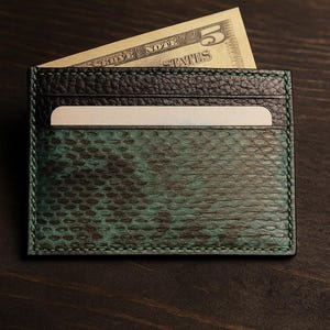 Könnte beinhalten: Ein grüner und schwarzer Kartenhalter aus Leder mit Schlangenmuster. Eine Fünf-Dollar-Note ist teilweise im Kartenhalter sichtbar. Der Kartenhalter liegt auf einer dunklen Holzoberfläche.