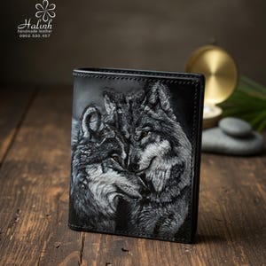 Könnte beinhalten: Schwarzes Lederportemonnaie mit einem Graustufenbild von zwei Wölfen. Das Portemonnaie zeigt ein geprägtes Wolfsdesign und den Text "halinh handmade leather". Das Portemonnaie wird auf einer Holzoberfläche präsentiert.