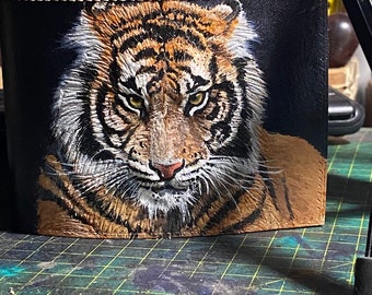 May include: Black leather wallet featuring a detailed painting of a tiger's face. The tiger has orange and black stripes, white fur, and piercing yellow eyes. The wallet is stitched around the edges.