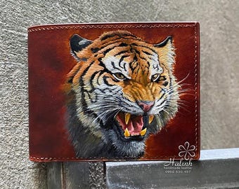 May include: Brown leather wallet featuring a detailed, hand-painted tiger face with orange, black, and white fur. The tiger is snarling, with visible teeth. The wallet has a stitched border and a dark red background.