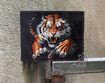 May include: Black leather wallet featuring a detailed, hand-painted tiger design. The tiger is depicted with orange and black stripes, white fur, and an open mouth, set against a dark background. The wallet has a classic bi-fold design.