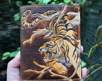 May include: A brown leather wallet featuring a detailed, hand-tooled relief carving of a tiger with black stripes, baring its teeth among tree branches. The leather has a burnished finish with visible stitching along the edges of the rectangular fold.