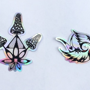 May include: Two holographic stickers. The first sticker features a black and white illustration of three mushrooms with a geometric design and a flower. The second sticker features a black and white illustration of a bird with a red heart.