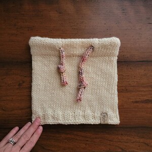 May include: A cream-colored knitted neck warmer with two pink and purple drawstrings. The neck warmer has a small brown label with the word "Willa" on it.
