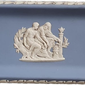 Könnte beinhalten: Eine blaue und weiße Keramik-Schmuckschale mit einem Reliefdesign von zwei Figuren in einer klassischen Umgebung. Die Schale hat einen dekorativen Rand.