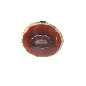 May include: A decorative agate bottle stopper with a polished, reddish-brown surface and a smaller, concentric agate slice at the center. The outer edge has a lighter, natural-looking border. The stopper is designed to fit a bottle.