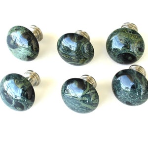 May include: Six oval-shaped cabinet knobs with a polished, speckled green and black stone appearance. Each knob has a silver-colored metal base. The knobs are arranged on a white background.