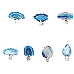 May include: A collection of seven lamp finials, each featuring a unique slice of blue agate stone mounted on a polished silver-toned metal base. The agate slices display various shades of blue, white, and translucent crystalline centers.