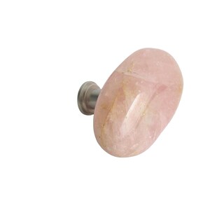 May include: A rose quartz cabinet knob with a polished, oval shape. The knob is a pale pink color with natural variations and a silver-toned metal base. This decorative hardware is suitable for drawers and cabinets.