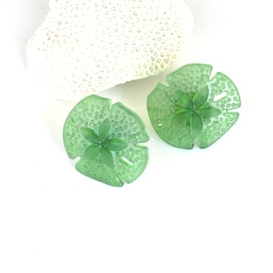 May include: A pair of green sand dollar earrings with a raised flower design in the center. The earrings are made of a translucent material and have a textured surface.