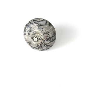 May include: A round, decorative stone with a marbled pattern in shades of gray and white. The stone has a smooth, polished surface and is set against a white background. The stone is a decorative item.