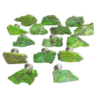 May include: A collection of sixteen green stone cabinet knobs with silver-toned metal bases. The knobs have a natural, rough-hewn appearance, with variations in color and texture. The knobs are arranged against a white background.