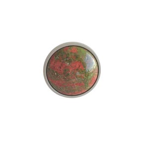 May include: A circular cabochon featuring a polished, mottled surface with a blend of deep red and moss green mineral inclusions, encased within a smooth, thin, silver-toned metal frame that creates a clean, defined border around the stone.