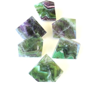 May include: A collection of six green and purple fluorite crystals, each with a unique shape and pattern. The crystals are arranged on a white surface.