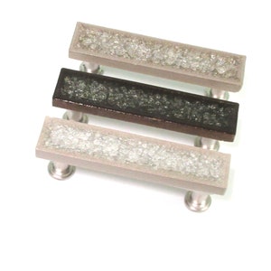 May include: Three rectangular cabinet pulls with a brushed metal finish and a decorative inset. The inset is filled with crushed glass in shades of clear, gray, and black. The pulls are mounted on a white background.
