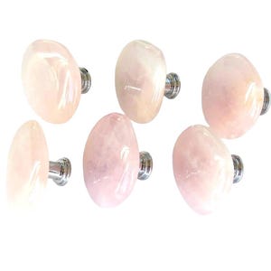 May include: A set of six cabinet knobs featuring polished, rounded, pale pink rose quartz stone faces attached to circular silver-toned metal bases. Each knob has a smooth, translucent surface with natural variations in the light pink stone.