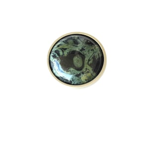 May include: A round, decorative cabinet knob with a gold-colored rim. The center features a swirling pattern of dark green, black, and gold colors. The knob is set against a plain white background.
