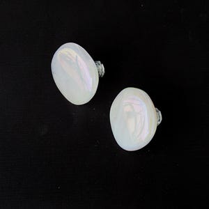 May include: Two white, oval-shaped stud earrings with iridescent surfaces. The earrings have silver-colored posts and are set against a black background. The earrings are a jewelry item.