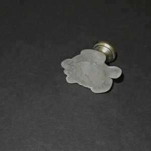 May include: A white glass cabinet knob shaped like a turtle with a silver base.