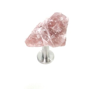 May include: A rose quartz cabinet knob with a polished stainless steel base. The pink stone has a rough, natural texture. The knob is designed for furniture and adds a touch of natural elegance.