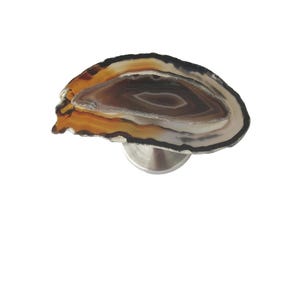 May include: A decorative cabinet knob featuring a polished agate slice. The agate displays concentric bands of brown, orange, and white, mounted on a silver-toned metal base. This unique hardware adds a natural touch.