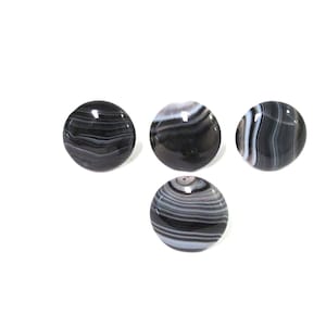 May include: Four polished, round stones with a banded pattern of black, white, and gray. The stones have a glossy finish and are arranged against a white background. The stones appear to be agate.