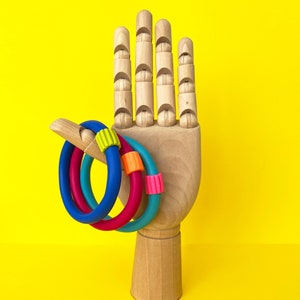 Pode incluir: Um manequim de mão de madeira exibe quatro pulseiras de silicone coloridas. As pulseiras são azuis, verde-azulado, rosa e uma combinação de rosa e laranja. O fundo é amarelo brilhante.