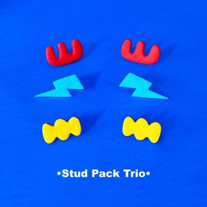 May include: A set of six colorful stud earrings. The earrings are shaped like lightning bolts, zigzags, and wavy lines. The earrings are red, yellow, and blue. The text "Stud Pack Trio." is printed below the earrings.