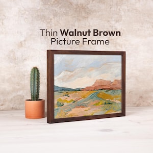Op de afbeelding: Een ingelijst landschapsschilderij met een dunne walnootbruine lijst. Het kunstwerk toont een woestijnlandschap met een weg, bergen en een bewolkte lucht. Links van het frame staat een cactus in een pot. Tekst op de afbeelding: Thin Walnut Brown Picture Frame.