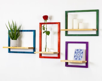 May include: Four colorful wooden shelves with a teal, red, purple, and green frame. The shelves are mounted on a white wall. The top shelf has two white candles and a third candle that is partially visible. The second shelf has a single red rose in a glass vase. The third shelf is empty. The bottom shelf has a small blue and white tile.
