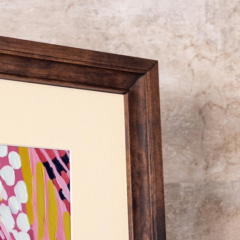Detailed corner close-up of a dark solid birch wood picture frame, highlighting the rich, natural wood grain. The frame includes a high-quality custom beige mat board displaying vibrant pink abstract art, showcasing the premium craftsmanship of this custom wood frame.