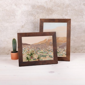 Op de afbeelding: Twee ingelijste landschapskunstprints met donkerbruine houten frames. Het kunstwerk toont een woestijnlandschap met bergen, cactussen en een gedempte kleurenpalet van beige, groen en bruin. Links een kleine cactus in een oranje pot.