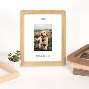 May include: A light-colored wooden picture frame containing a white mat with the name Milo printed above a photograph of a yellow Labrador retriever in a field, and the years 2014-2026 printed below the photograph in a dark brown font.