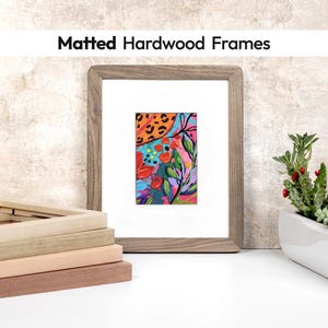 May include: A matted hardwood frame with a colorful abstract painting. The painting features red, blue, and pink floral elements with a leopard print accent. The text "Matted Hardwood Frames" is at the top. Several wood frames are stacked in the foreground.