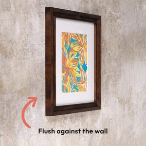 Side profile of a dark solid birch wood picture frame hanging completely flush against a textured wall. Highlights the sleek, low-profile design of this hanging photo frame with a custom white mat.