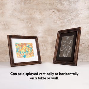 Two solid wooden photo frames leaning on a table, demonstrating vertical portrait and horizontal landscape display options. The custom wood frames feature white and black custom matting displaying colorful and monochrome art.