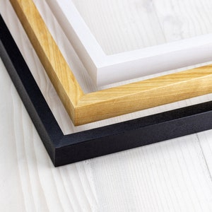 Könnte beinhalten: Drei Bilderrahmen, einer schwarz, einer aus Naturholz und einer weiß, sind übereinander gestapelt. Die Rahmen sind alle rechteckig und haben ein schlichtes Design.