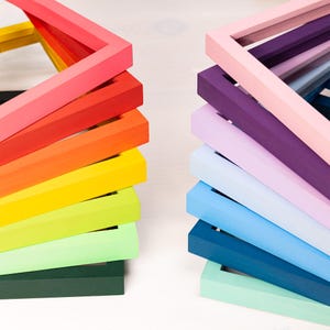Puede incluir: Dos pilas de marcos de fotos cuadrados de madera en un espectro de colores. La pila izquierda presenta tonos cálidos que incluyen rojo, naranja, amarillo y verde lima. La pila derecha presenta tonos fríos que incluyen rosa, morado, lavanda, azul y verde menta.