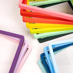 以下が含まれることがあります： ピンク、赤、オレンジ、黄色、ライトグリーン、ダークグリーン、紫、ラベンダー、水色、濃い青、ティールなど、さまざまな無地の長方形のフォトフレームのコレクション。それらが重なり合うように積み重ねられて配置されています。