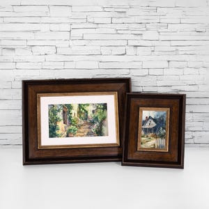 Pode incluir: Duas pinturas de aquarela emolduradas. A pintura maior representa um caminho de pedra que leva a uma colina com vegetação exuberante e um edifício ao fundo. A pintura menor mostra uma pequena casa com uma cerca branca de madeira e um caminho que leva a ela.