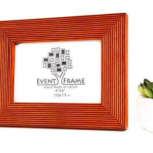 以下が含まれることがあります： 溝のデザインが施された、オレンジ色の木製の4x6インチの写真フレーム。このフレームはラトビア製で、前面に「EVENT FRAME」と「HAND MADE IN LATVIA 4"x"6" 10x15 cm」の文字が印刷されています。