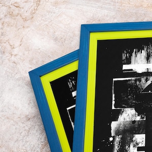May include: Two framed art prints with a modern abstract design. The frames have a blue exterior and a bright yellow-green interior. The artwork is black and white, with a textured, graphic style. The frames are displayed on a textured, neutral background.