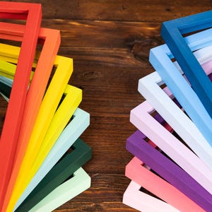 May include: A collection of colorful wooden picture frames in a variety of hues, including red, orange, yellow, green, blue, purple, and pink. The frames are arranged in two separate fan-like formations against a dark wood background.