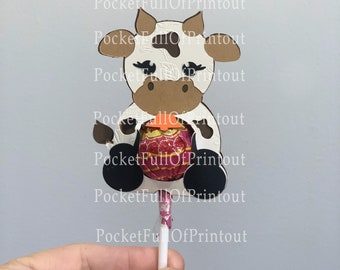 May include: A white and brown cow lollipop holder with black eyes and a pink and white lollipop inside. The holder is attached to a white stick.