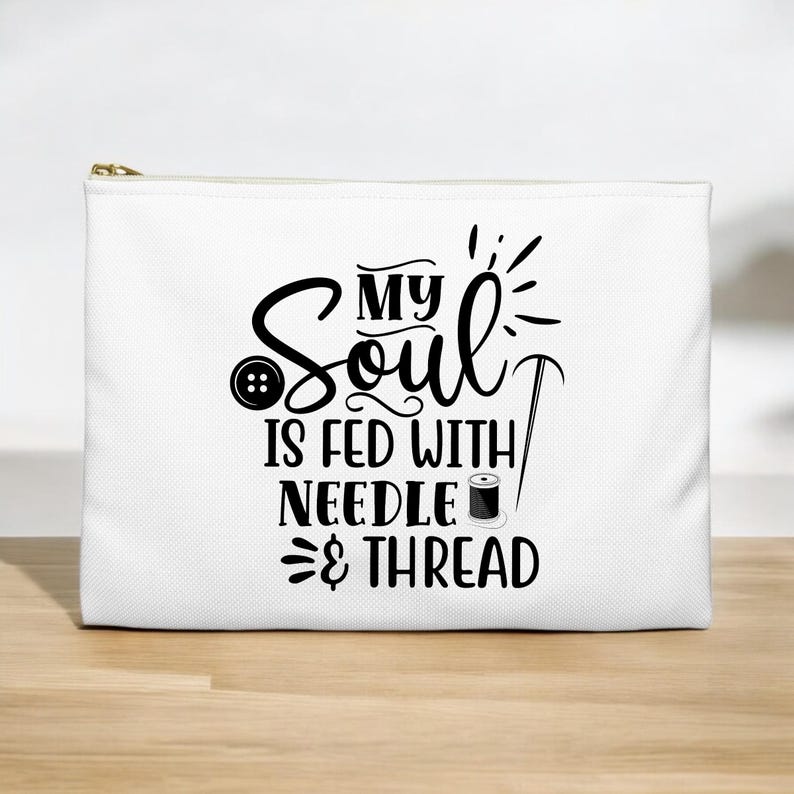 以下が含まれることがあります： 黒い文字で「My Soul is Fed with Needle & Thread」と書かれた白いジッパー付きポーチ。テキストは針、糸、ボタンでスタイリッシュにデザインされています。